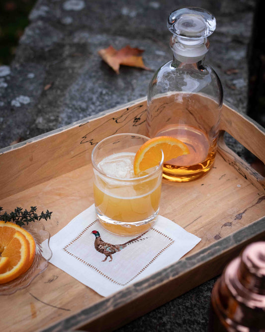 Bourbon sour with a slice of orange sitting on a linen cocktail napkin embroidered with a pheasant, sitting on a wooden tray.