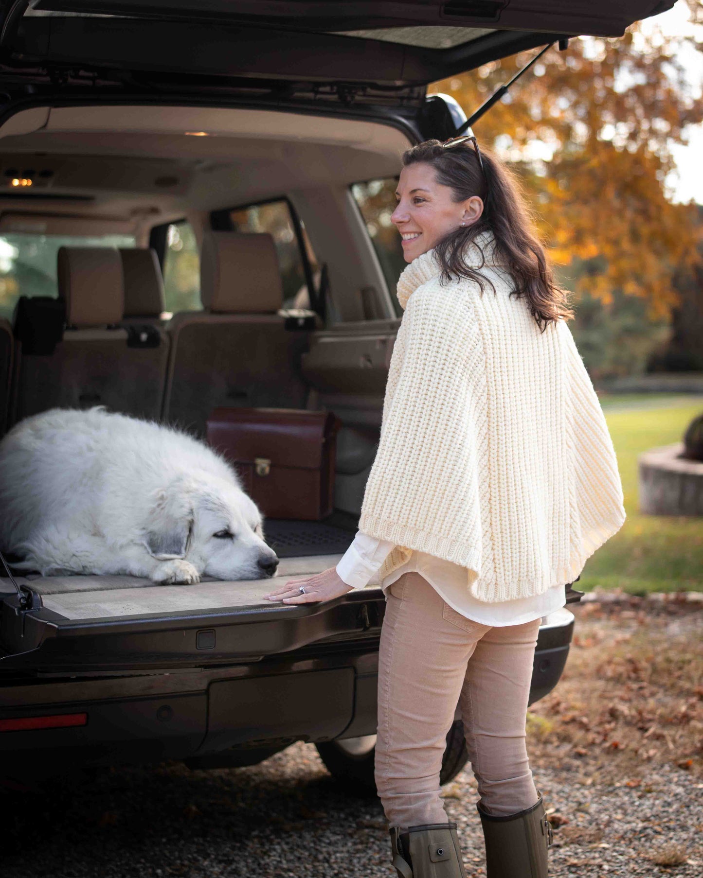 Woman standing next to a white dog in an open car trunk, with a scenic background.