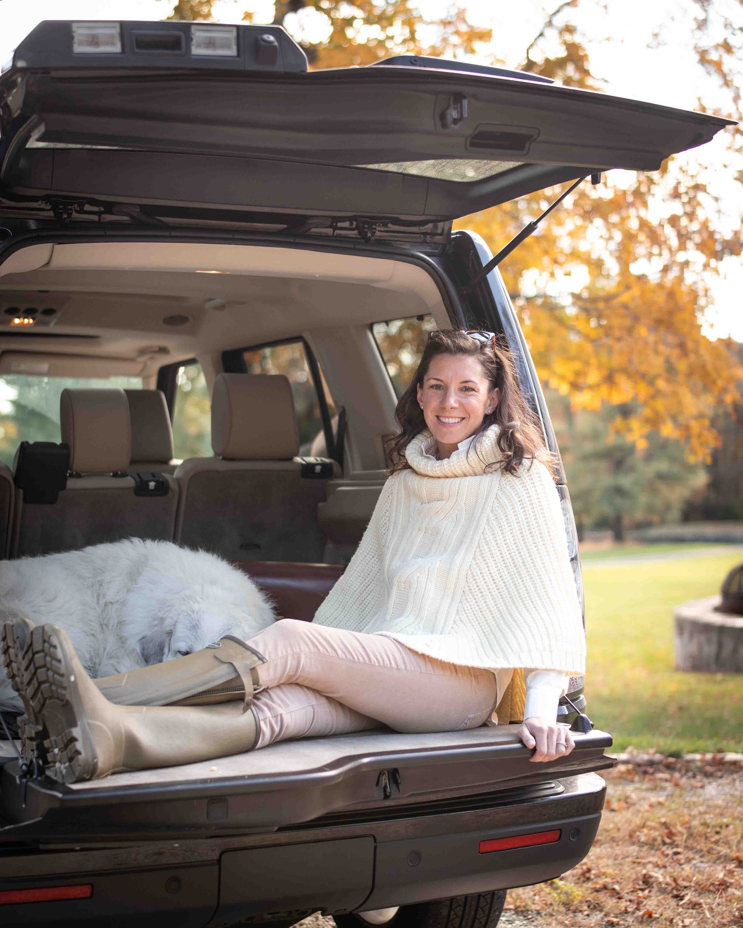 Woman sitting in the open trunk of a car wearing a cream poncho sweater, with a dog at her side , surrounded by autumn foliage.