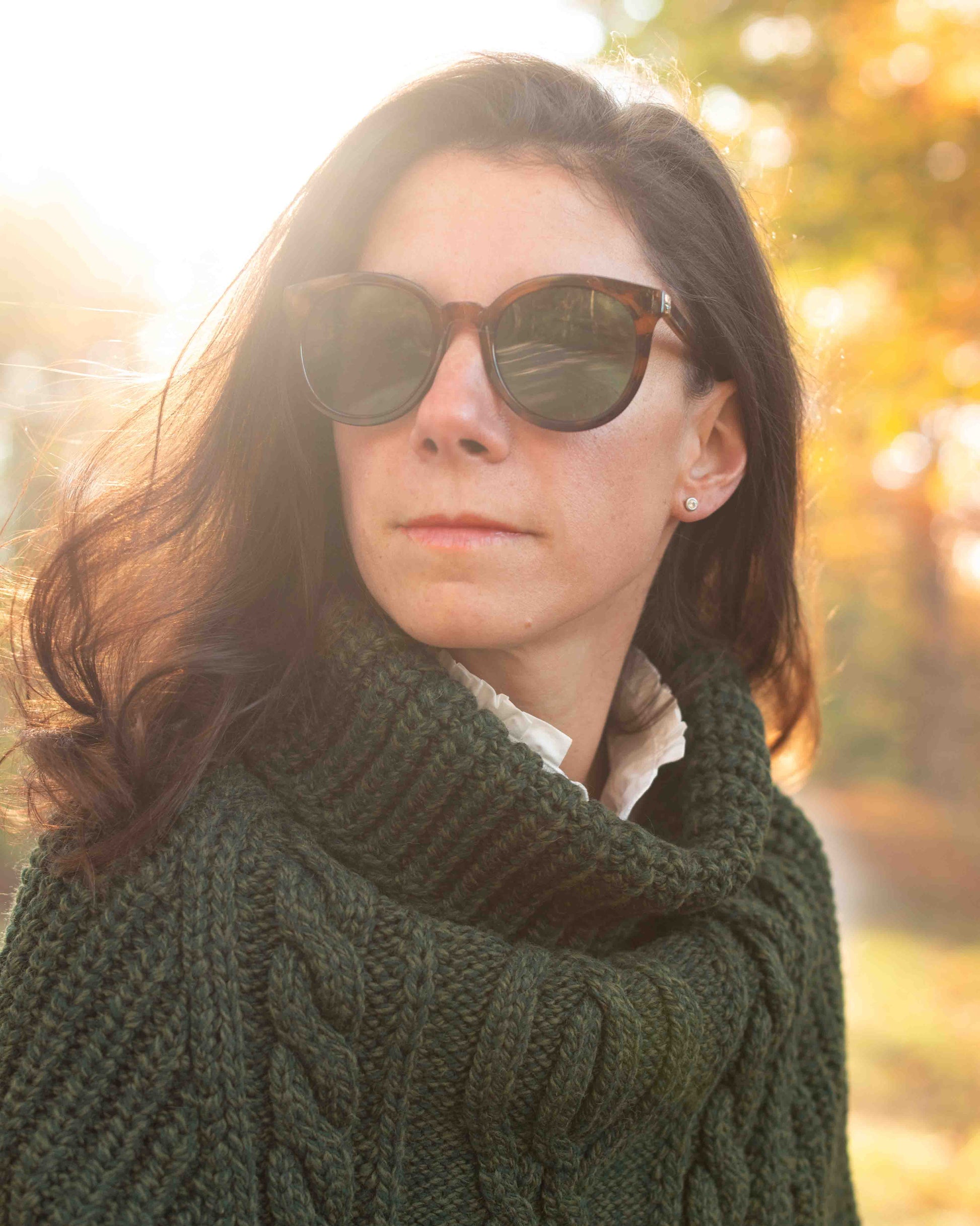 Woman wearing sunglasses and a green sweater with a blurred autumn background
