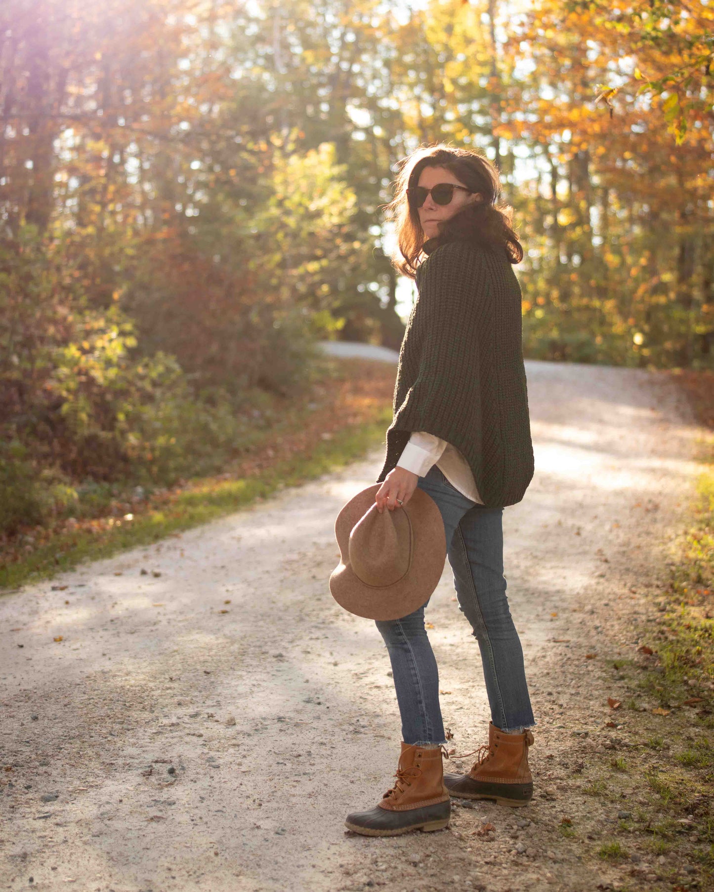Woman standing on a dirt road in a forest during autumn, wearing a wool poncho, holding a hat.